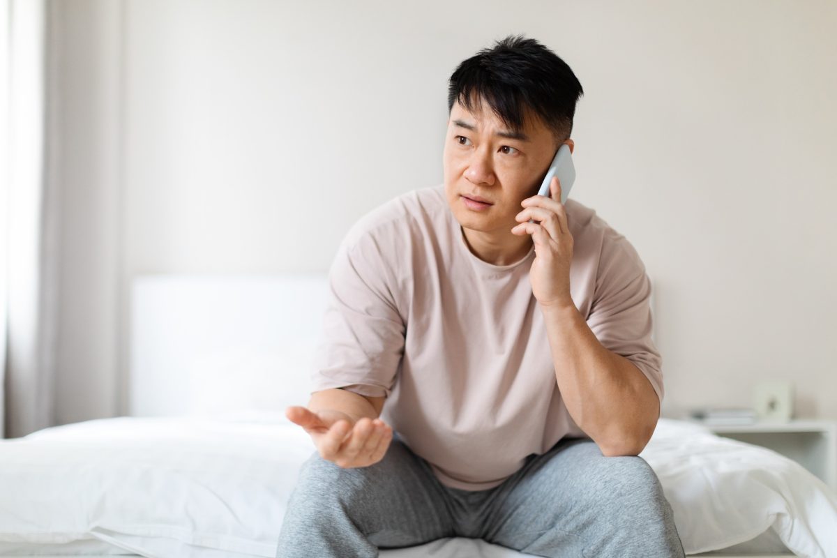 Puzzled chinese man sitting on bed, have phone call
